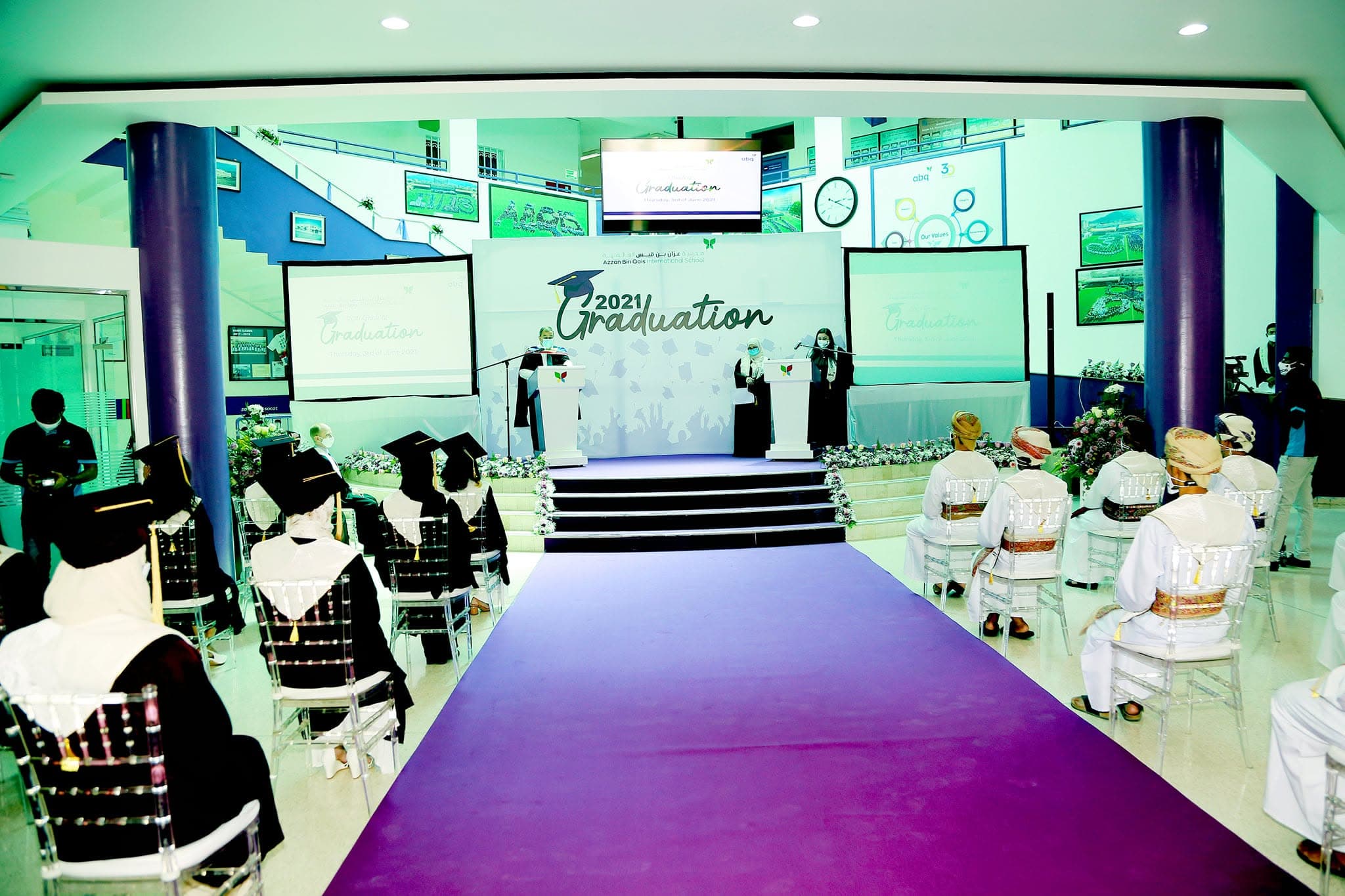 Graduates in academic gowns and traditional Omani attire sit facing a 2021 graduation stage.