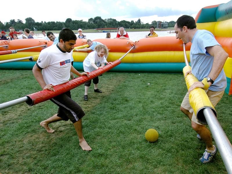 People playing human foosball in an inflatable arena on grass with a yellow ball.
