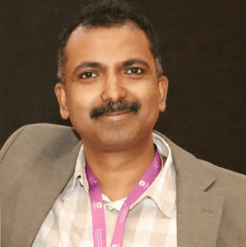 Smiling man with a mustache wearing a tan blazer and purple lanyard against dark background.