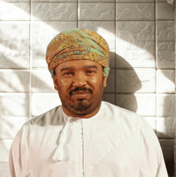 Man in a patterned Omani turban and white dishdasha posing against a white tiled wall.