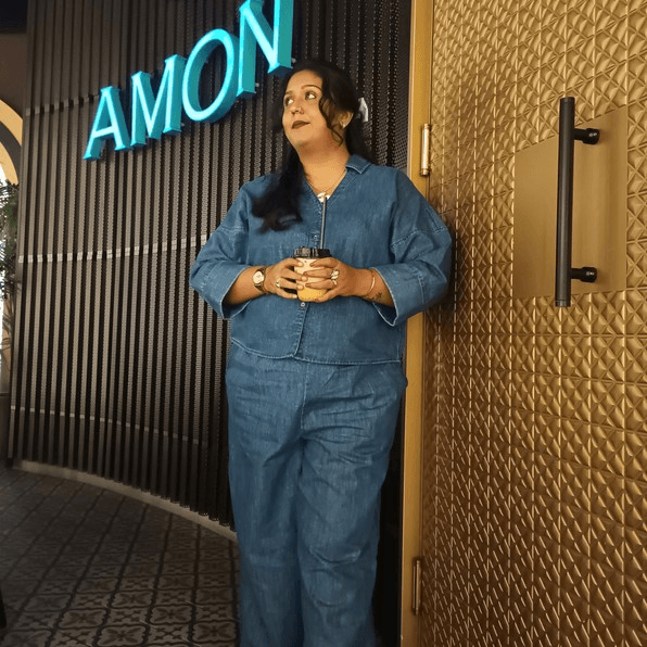 Woman in blue denim holding a drink near a neon sign and textured gold door.